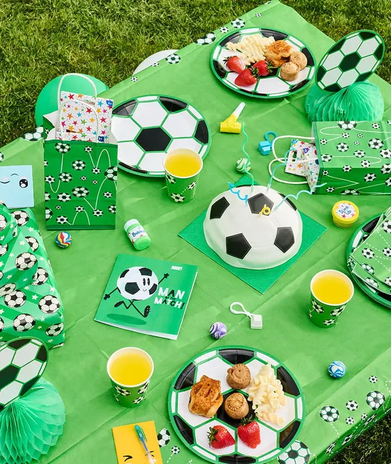 Football-themed party picnic with football-print tableware, a football-shaped cake, party bags, snacks, drinks, and green party decorations on the grass. Football-themed party picnic with football-print tableware, a football-shaped cake, party bags, snacks, drinks, and green party decorations on the grass.